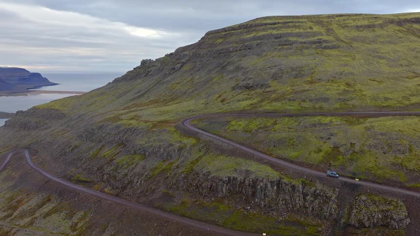 Aerial view of a camper van driving along a winding dirt road on a vast, rugged mountainside overlooking the ocean