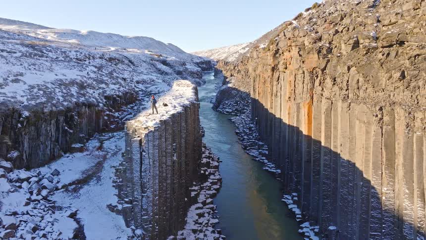 An adventurous person stands triumphantly atop the magnificent, snow-covered basalt columns of Studlagil Canyon in Iceland as the aerial camera pulls back to reveal the epic winter landscape