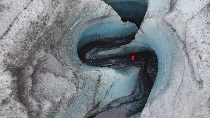 An aerial view descends into a winding, blue-ice canyon on a glacier, revealing a lone person in a red jacket standing in the meltwater