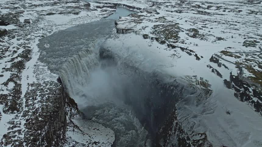 A stunning aerial view reveals a powerful waterfall crashing into a deep canyon amidst a vast, desolate, snow-covered winter landscape