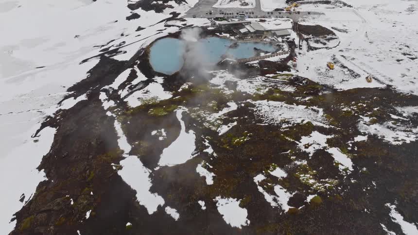 A stunning high-angle aerial shot rises over a snowy volcanic hill to reveal the steaming blue waters of the Myvatn Nature Baths in Iceland