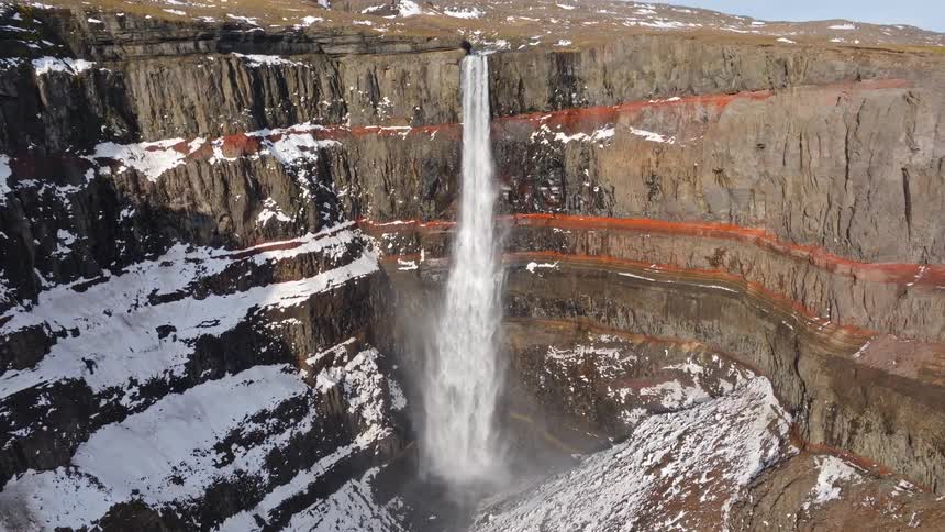 A majestic aerial reveal of the Hengifoss waterfall plunging down a snow-dusted cliff face with striking red sediment layers in Iceland