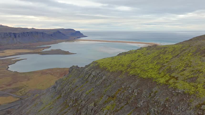 A slow aerial pullback reveals a vast, moss-covered mountainside overlooking a dramatic Icelandic fjord and the distant ocean