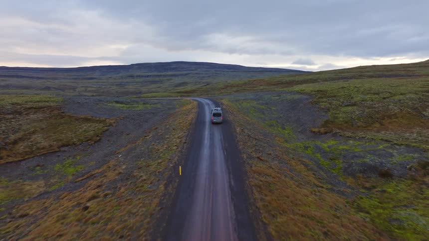 An aerial view follows a camper van driving along a remote, winding dirt road through the vast, mossy highlands of Iceland