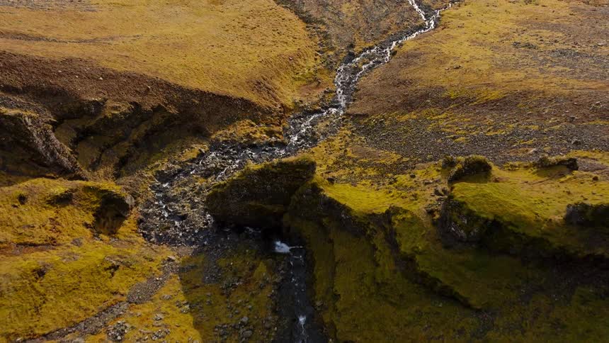Top-down aerial view slowly pulls back from a waterfall in a mossy canyon to reveal a vast, rugged landscape and the sea