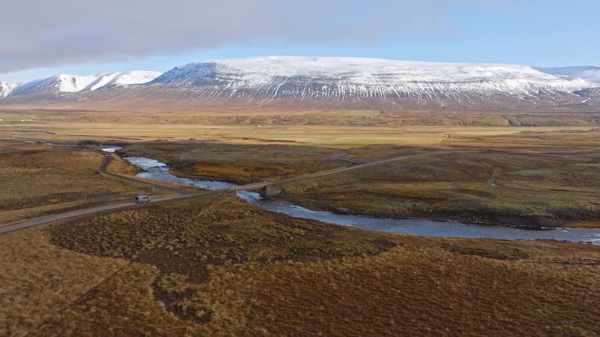 Aerial footage tracks a lone camper van on a road trip through a vast Icelandic landscape as it crosses a bridge over a river with snow-capped mountains in the background
