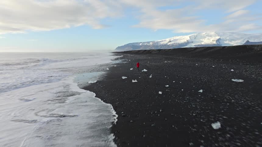 A dramatic aerial pullback reveals a person in a red coat walking along Iceland's famous Diamond Beach, surrounded by black sand, ocean waves, and a distant glacier