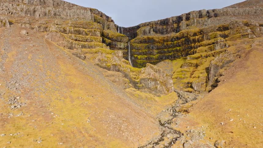 An aerial drone shot slowly pushes towards a majestic waterfall cascading down a tiered, moss-covered cliff in a rugged golden valley