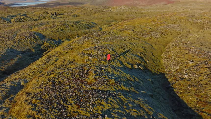 Aerial view reveals a person in a red coat standing on a vast mossy hill in a stunning Icelandic volcanic landscape at sunrise