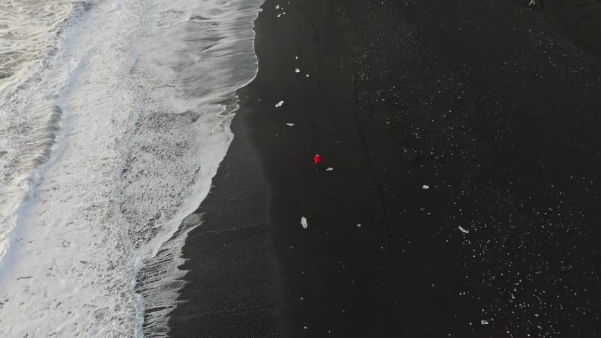 A dramatic top-down aerial view of a person in a red jacket on a black sand beach pulls back to reveal the vast, icy coastline of Iceland