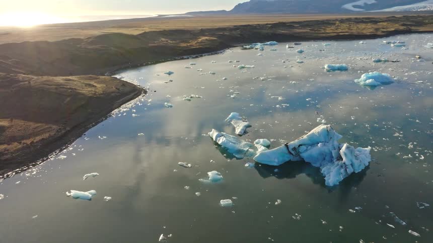 A majestic aerial view captures icebergs floating in a calm glacier lagoon in Iceland as the sun sets over the distant ocean