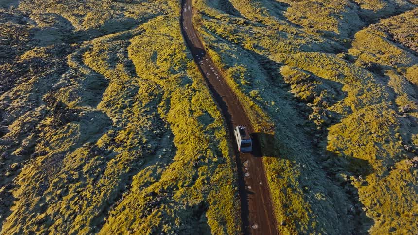 An epic aerial shot follows a van driving through Iceland's stunning green moss-covered lava fields during a golden sunrise