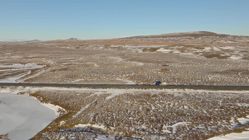 An aerial view of two cars driving on a remote road that cuts through a vast, snowy, and barren landscape under a clear sky