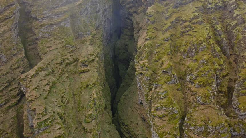 Aerial view tilting down into a deep, narrow canyon with steep, moss-covered rock walls and misty clouds