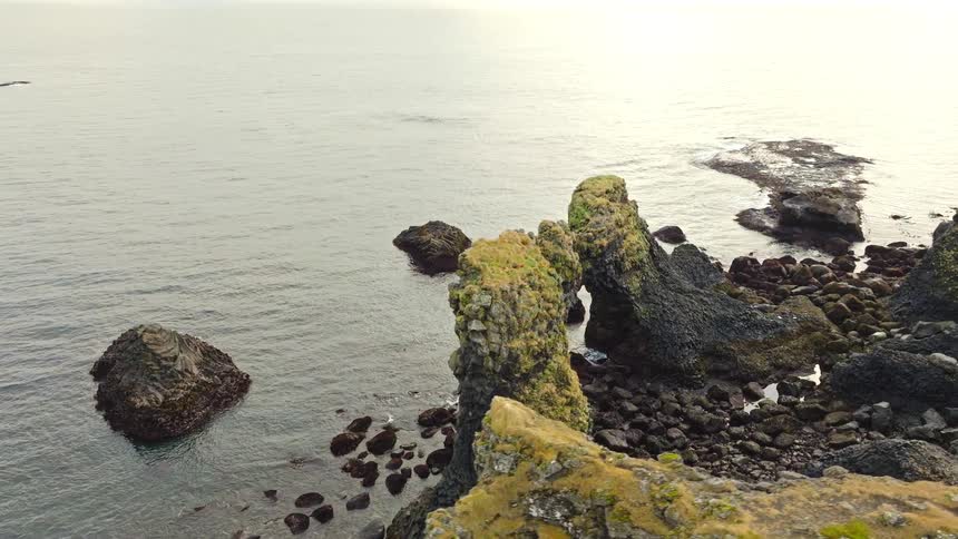 Aerial drone footage slowly reveals the stunning moss-covered Gatklettur rock arch formation along the tranquil Icelandic coastline
