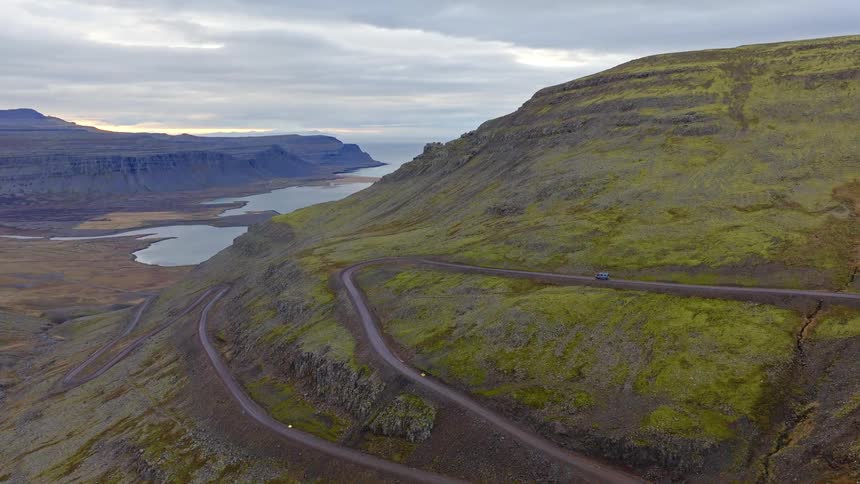 An aerial view shows a lone camper van driving along a winding gravel road through the dramatic, mossy mountains of the Icelandic Westfjords