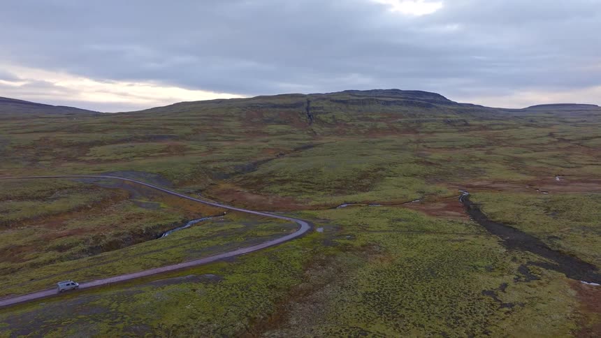 An aerial drone shot ascends over a camper van parked on a winding road through Iceland's vast, green, moss-covered highlands