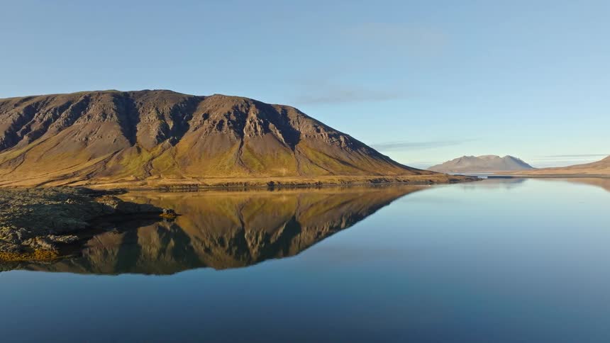 Drone flies backward over a tranquil fjord in Iceland, revealing a perfect mirror reflection of a majestic mountain range on the calm water