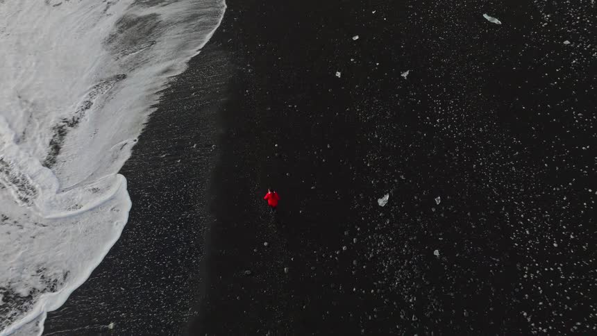 Top-down aerial shot tilts up to follow a solitary person in a red coat walking along the dramatic black sand shoreline of Diamond Beach in Iceland