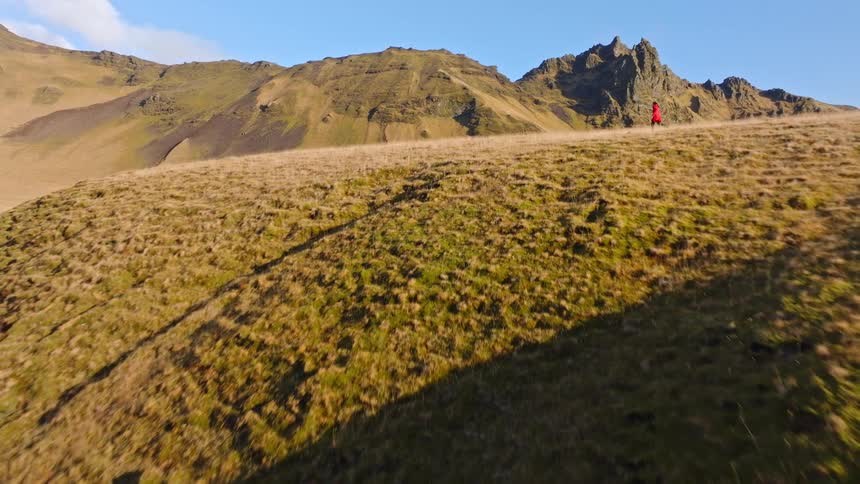 Aerial footage tracks a woman in a bright red coat as she runs across a vast and scenic mountain landscape during golden hour
