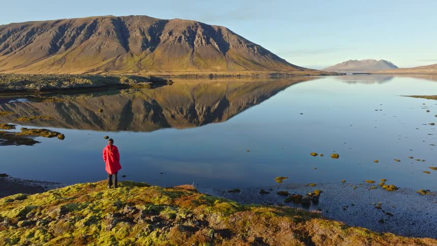 A woman in a red coat stands on a mossy shore, gazing at a majestic mountain perfectly reflected in the still water