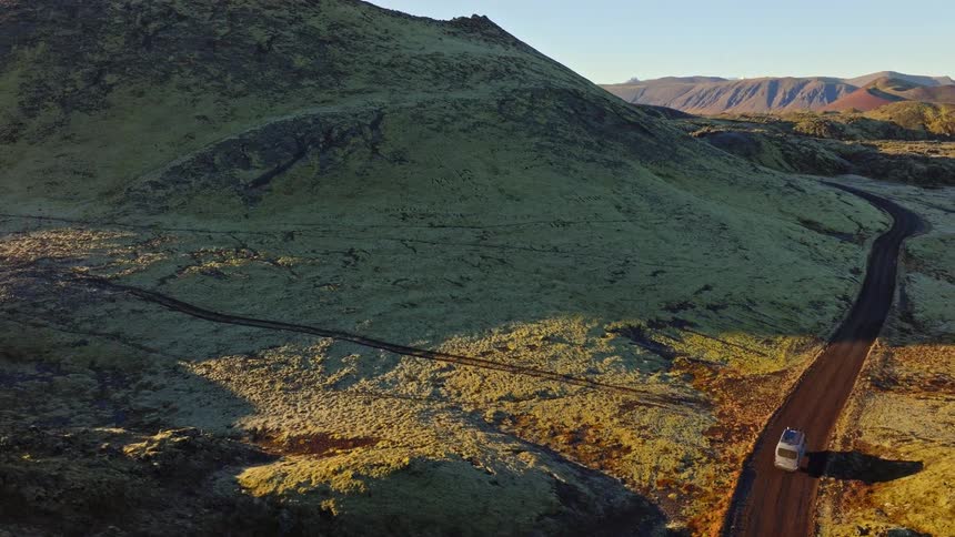 An epic aerial shot follows a camper van driving on a remote dirt road, rising to reveal the vast, moss-covered volcanic landscape of Iceland