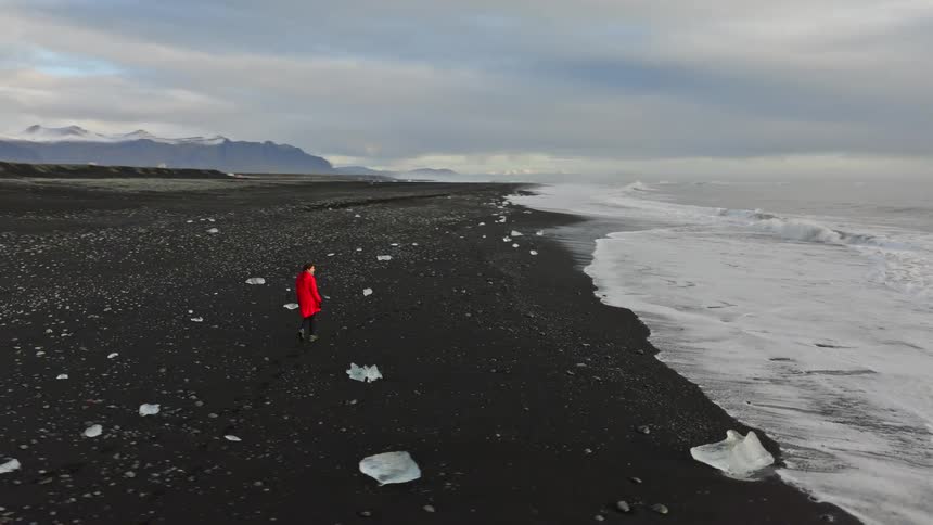 An aerial view shows a person in a red coat walking along the dramatic black sand beach in Iceland, surrounded by ice chunks, crashing waves, and snow-capped mountains