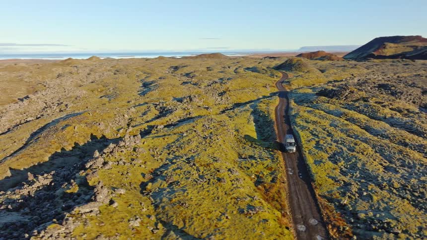 An aerial shot follows a recreational vehicle as it travels along a remote road through a stunning moss-covered volcanic landscape at golden hour