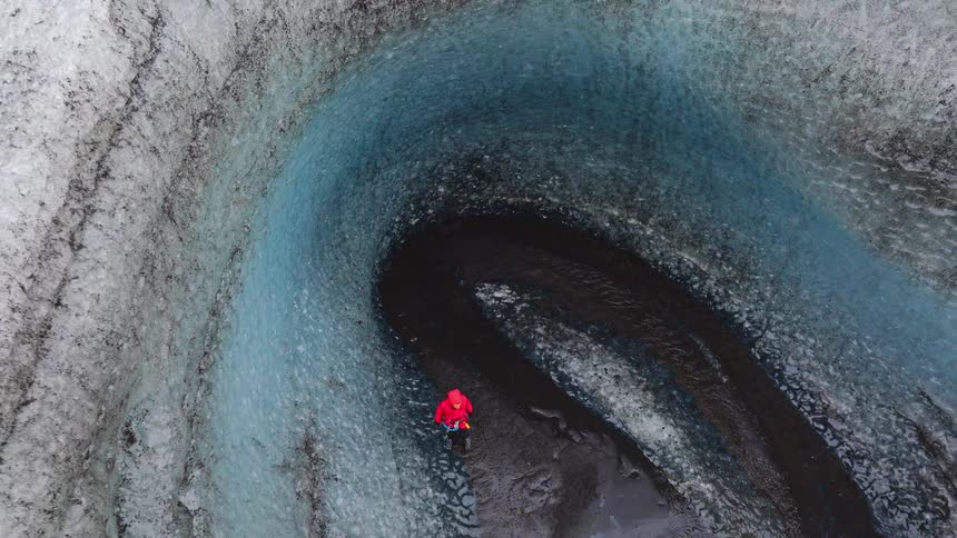 A high-angle aerial shot pulls back from a person in a red jacket standing inside a majestic blue ice glacier, revealing its vast and winding structure