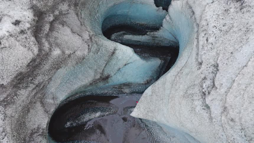High-angle aerial shot reveals a person in a red jacket standing in a stunning S-shaped ice canyon as the camera ascends