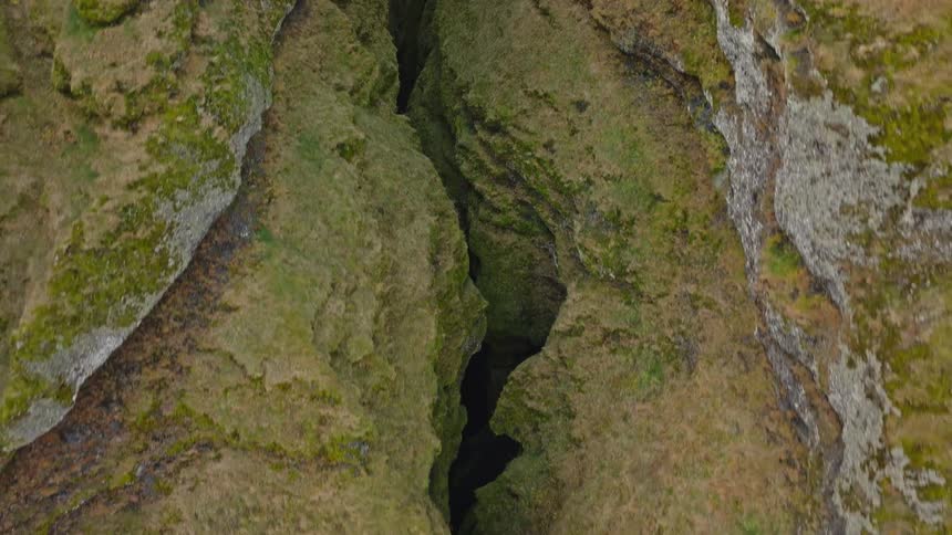 A drone flies backward out of a deep, green, mossy canyon, tilting up to reveal the magnificent and rugged mountainscape