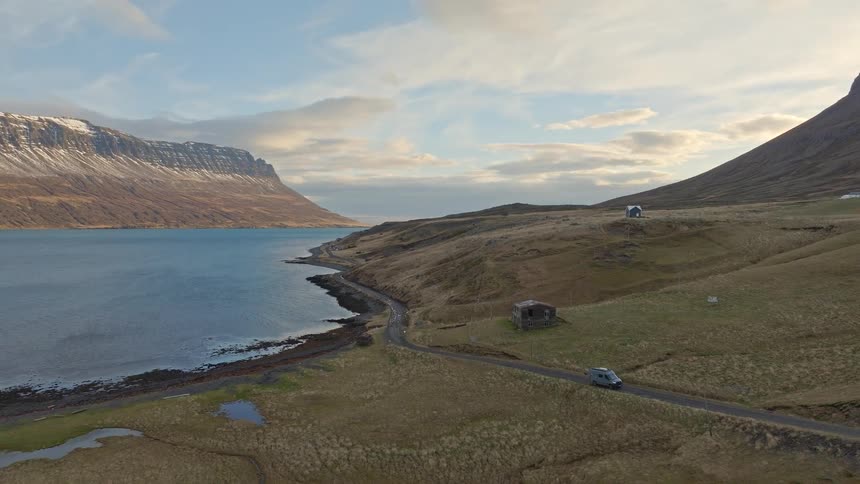 Aerial view of a camper van on a scenic drive along a remote coastal road next to a vast fjord and snow-capped mountains in Iceland