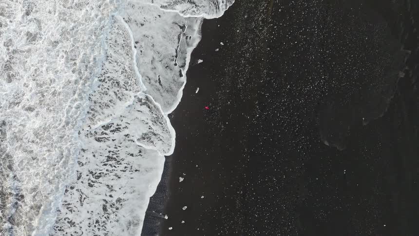 A striking aerial top-down view shows a person in a red jacket lying on a vast black sand beach as white ocean waves crash ashore amongst scattered pieces of ice