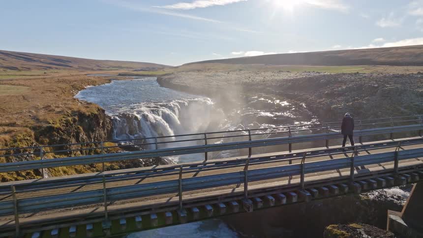 Drone pulls back from a powerful Icelandic waterfall to reveal a lone traveler walking across a bridge on a sunny day