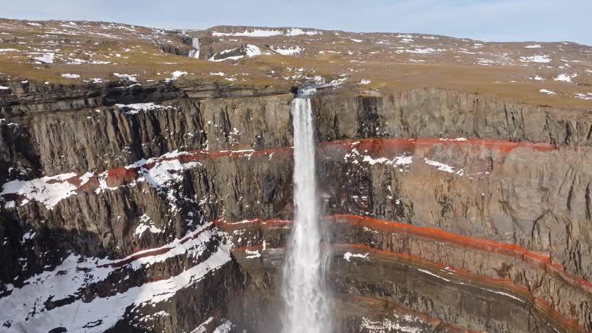 Aerial drone footage reveals Hengifoss waterfall cascading over a red-layered canyon in the vast, snowy highlands of Iceland