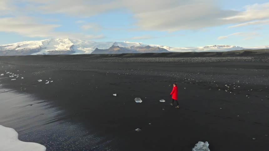 Aerial tracking shot follows a woman in a bright red coat walking along the famous black sand Diamond Beach in Iceland, with glaciers in the background