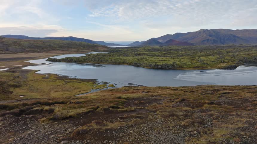An aerial drone shot follows a hiker in a bright red coat walking along a trail, revealing a stunning Icelandic landscape with a lake and volcanic mountains
