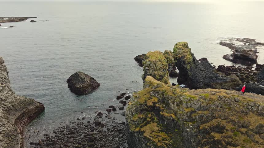 An aerial shot reveals a person in a red coat standing on a rugged Icelandic cliff, admiring the view of a massive natural rock arch over the ocean