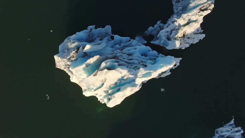 An aerial top-down shot slowly rises to reveal a magnificent, textured iceberg floating in a dark, tranquil glacial lagoon