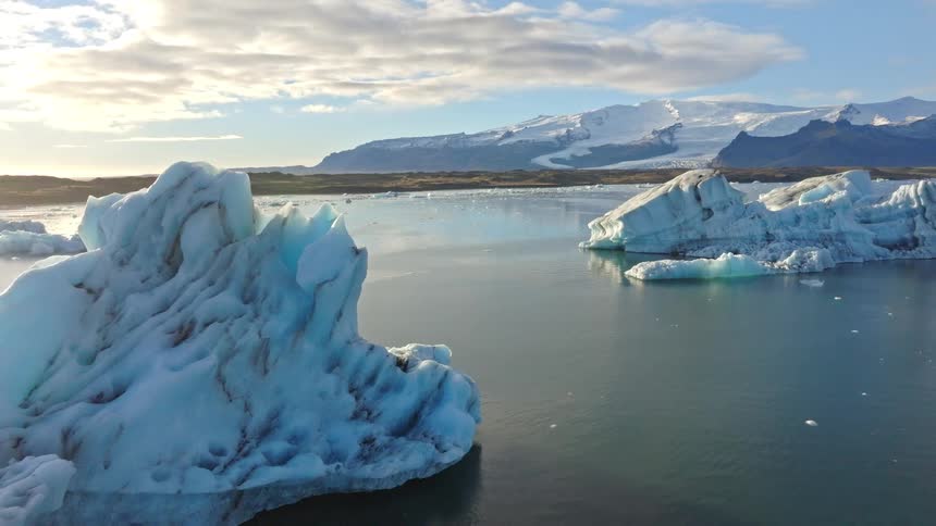 A slow aerial pullback reveals majestic icebergs floating in the serene Jokulsarlon glacier lagoon, with the Vatnajökull glacier in the background