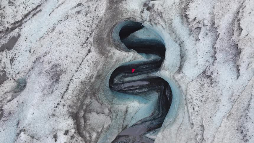 A dramatic top-down aerial shot zooms out from a person in red exploring a winding ice canyon carved by a meltwater river on a massive glacier
