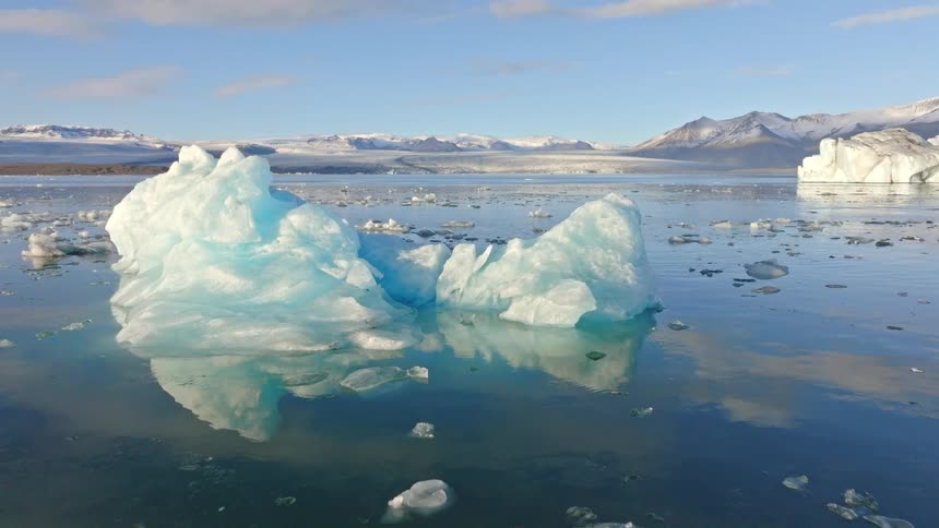 Aerial view pulling back over a majestic blue iceberg floating in a calm glacial lagoon in Iceland, with snow-covered mountains in the distance
