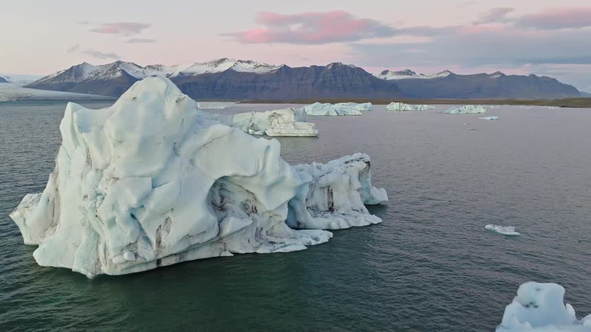 A serene aerial pull-back shot over a glacial lagoon in Iceland, revealing massive, sculpted icebergs against a backdrop of snow-capped mountains at sunset