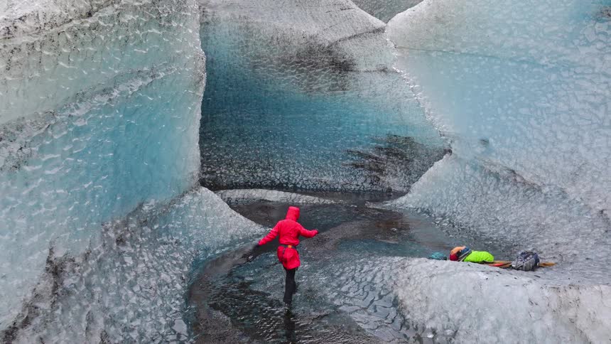 An aerial view follows a hiker in a red jacket as they explore a majestic blue ice canyon carved into a vast glacier