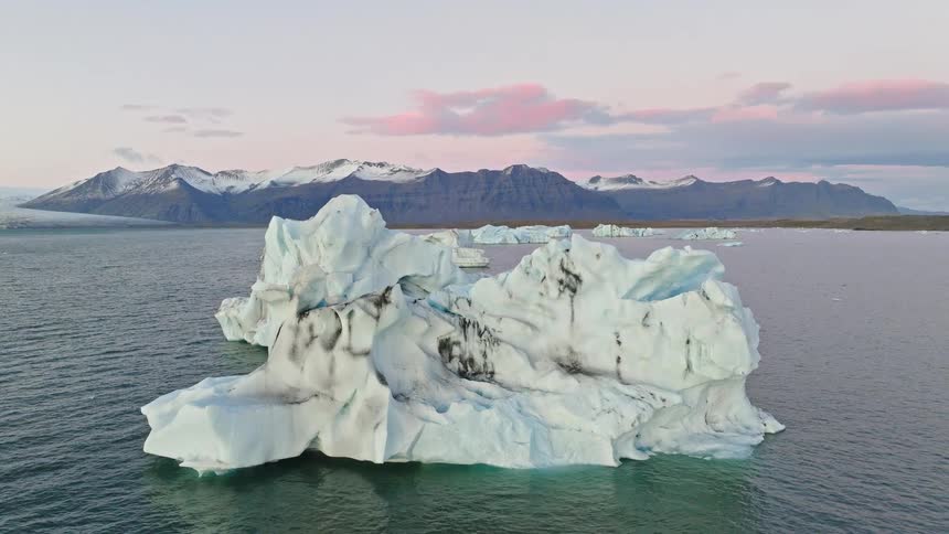 A static shot of a large iceberg floating in calm water pulls up to reveal a beautiful glacial lagoon with snow-capped mountains at sunset in Iceland