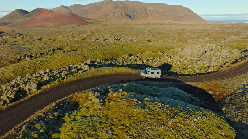 An aerial view follows a camper van driving along a remote, winding dirt road through a vast, moss-covered volcanic landscape at sunrise