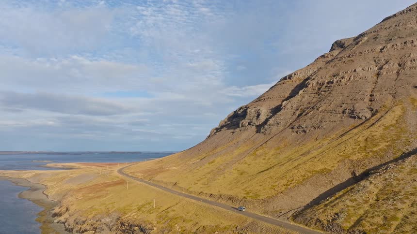 Aerial footage follows a car on a scenic road trip along a coastal highway at the base of a massive, rugged mountain in the Icelandic fjords