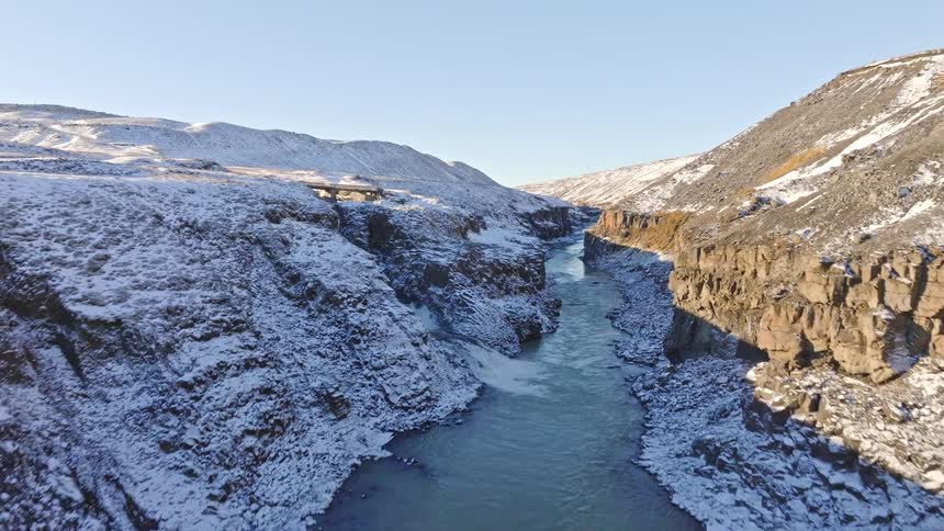 Majestic aerial flight through a stunning winter canyon in Iceland, revealing a rushing river and waterfall surrounded by snow-covered basalt columns