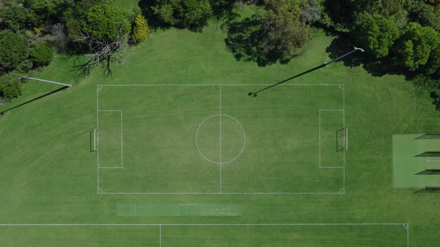 An aerial top-down view slowly ascends over an empty green soccer field surrounded by lush trees in a park