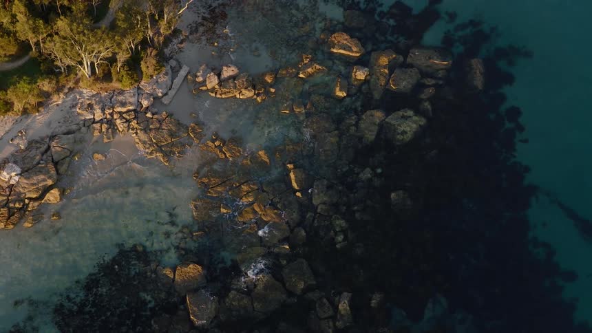 A stunning top-down aerial view slowly ascends over a rocky coastline with clear turquoise water during a peaceful sunrise